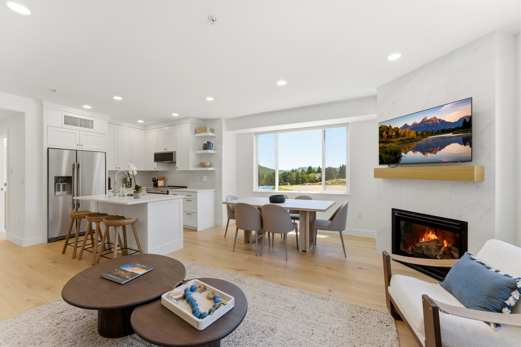 Open-concept living area featuring light wood-finish flooring, a contemporary fireplace with a stone surround, and large windows