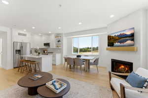 Open-concept living area featuring light wood-finish flooring, a contemporary fireplace with a stone surround, and large windows