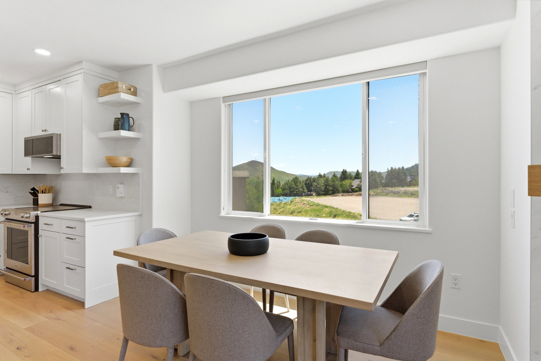 Contemporary kitchen and dining area featuring a large picture window, white shaker cabinetry, stainless steel appliances, and wood-finish flooring