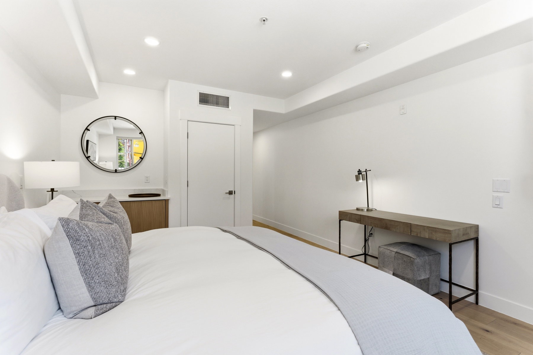 Recessed lighting illuminating the white walls, wood-finish flooring, built-in shelving with wood cabinetry, white door with black hardware, and a circular wall mirror with black framing