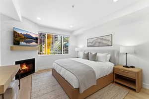 Contemporary bedroom featuring a built-in fireplace with a natural wood mantel, light wood-finish flooring, and a large window with roller shades