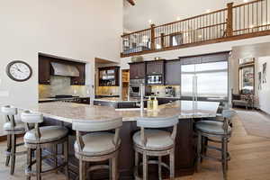 Kitchen featuring light wood finished floors, dark brown cabinets, light stone countertops, a towering ceiling, and tasteful backsplash