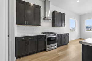 Kitchen with stainless steel gas range oven, wall chimney exhaust hood, light stone countertops, tasteful backsplash, and light wood-style flooring