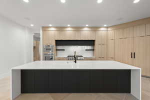 Kitchen with modern cabinets, light brown cabinetry, light stone counters, a large island with sink, and backsplash