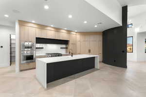 Kitchen with modern cabinets, an island with sink, light brown cabinetry, light stone countertops, and tasteful backsplash