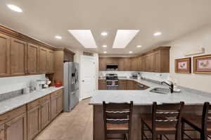 Kitchen with light stone countertops, a kitchen bar, a skylight, stainless steel appliances, and brown cabinets