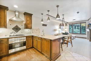 Kitchen with stainless steel oven, wall chimney exhaust hood, brown cabinetry, a peninsula, and light stone counters
