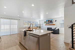 Kitchen with stainless steel dishwasher, wood finish floors, a glass covered fireplace, open floor plan, and a center island with sink