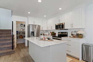 Kitchen with stainless steel appliances, white cabinets, an island with sink, light wood finished floors, and light stone counters