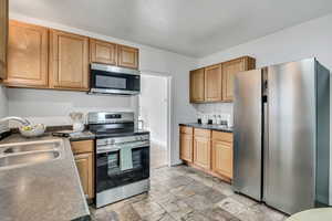 Kitchen with stainless steel appliances, dark countertops, light stone finish flooring, and brown cabinets