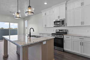Kitchen featuring appliances with stainless steel finishes, light stone countertops, a center island with sink, hanging light fixtures, and white cabinetry