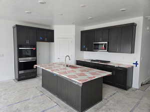 Kitchen with stainless steel appliances, a center island with sink, dark cabinetry, a textured ceiling, and light stone counters