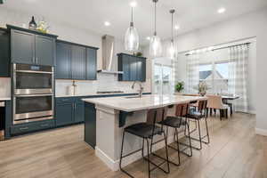 Kitchen featuring a breakfast bar area, backsplash, a kitchen island with sink, stainless steel double oven, and light wood finished floors
