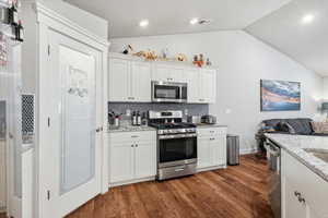 Kitchen featuring white cabinets, stainless steel appliances, lofted ceiling, light stone counters, and dark wood finished floors