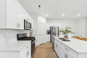 Kitchen featuring appliances with stainless steel finishes, white cabinets, light stone countertops, a center island with sink, and light wood-style flooring