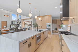 Kitchen featuring light brown cabinetry, light stone countertops, light wood-style flooring, range hood, and pendant lighting