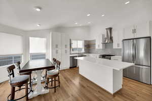 Kitchen featuring stainless steel appliances, white cabinetry, lofted ceiling, wall chimney exhaust hood, and light wood finished floors