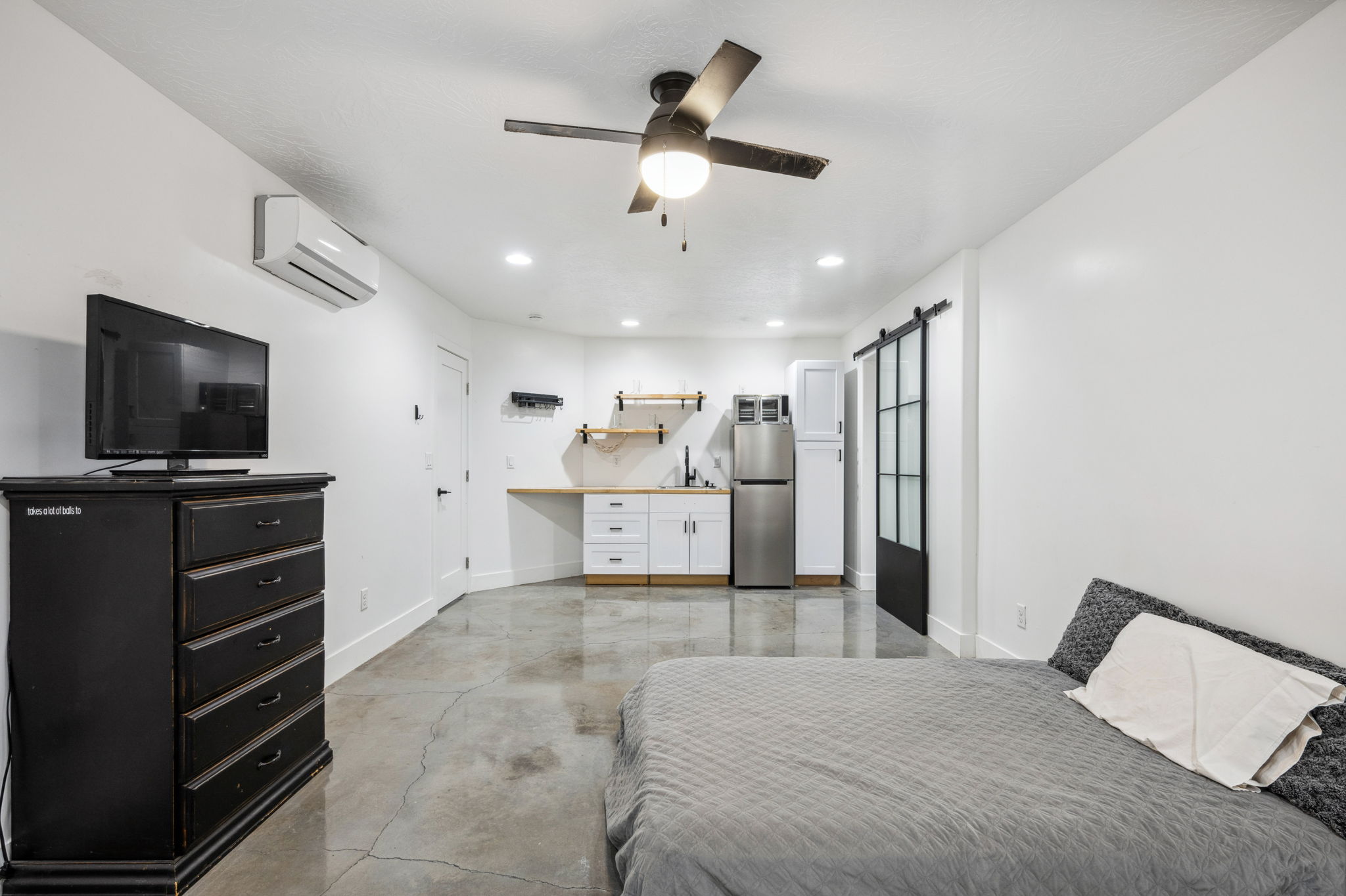 Bedroom with a barn door, finished concrete flooring, freestanding refrigerator, ceiling fan, and recessed lighting