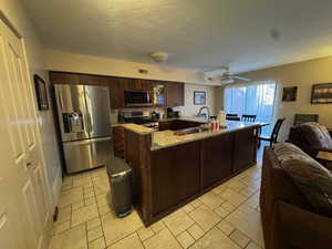 Kitchen with appliances with stainless steel finishes, light stone counters, open floor plan, dark brown cabinets, and ceiling fan