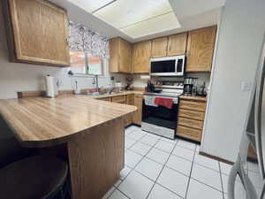 Kitchen featuring a peninsula, stainless steel appliances, a kitchen bar, brown cabinetry, and light tile patterned floors