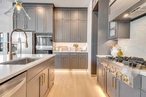 Kitchen with custom range hood, light wood-type flooring, appliances with stainless steel finishes, gray cabinets, and light stone countertops