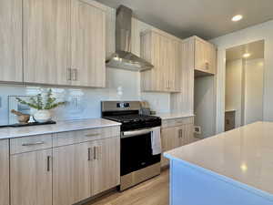 Kitchen with light brown cabinets, stainless steel gas range oven, wall chimney exhaust hood, modern cabinets, and light stone counters