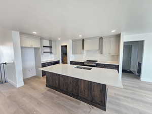 Kitchen featuring light stone countertops, a center island with sink, light wood finished floors, stainless steel gas range, and backsplash