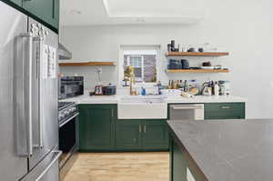 Kitchen with green cabinetry, stainless steel appliances, open shelves, light wood-type flooring, and wall chimney range hood