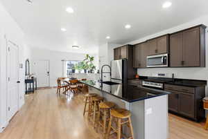 Kitchen featuring a kitchen island with sink, appliances with stainless steel finishes, light wood-type flooring, a kitchen breakfast bar, and dark stone counters
