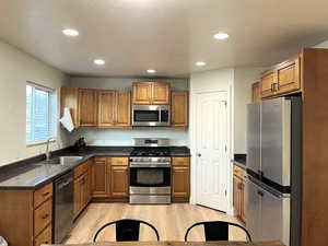 Kitchen with appliances with stainless steel finishes, brown cabinets, light wood-type flooring, recessed lighting, and dark stone counters