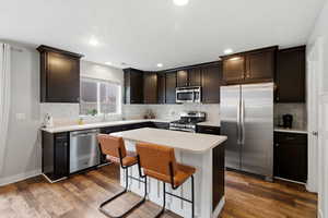 Kitchen with appliances with stainless steel finishes, dark wood finished floors, a center island, dark brown cabinetry, and a kitchen bar
