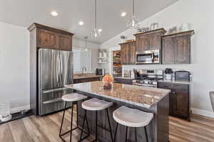 Kitchen featuring dark brown cabinetry, appliances with stainless steel finishes, light stone counters, a breakfast bar area, and a center island