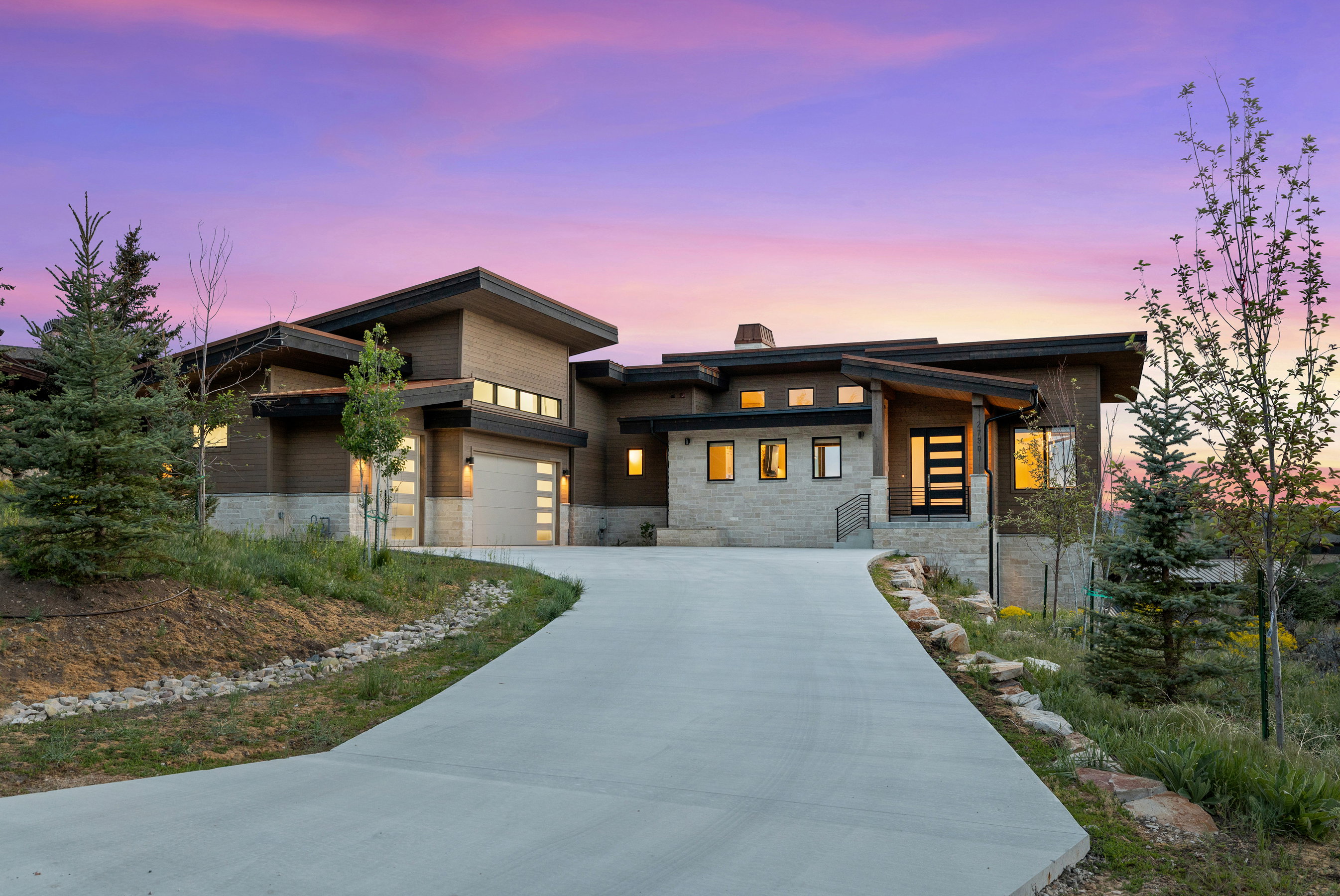 Contemporary architectural residence featuring a prominent concrete driveway, multi-toned exterior siding, an integrated two-car garage with modern doors, a distinctive front entry with a dark door and rectangular glass inserts, and surrounding natural la
