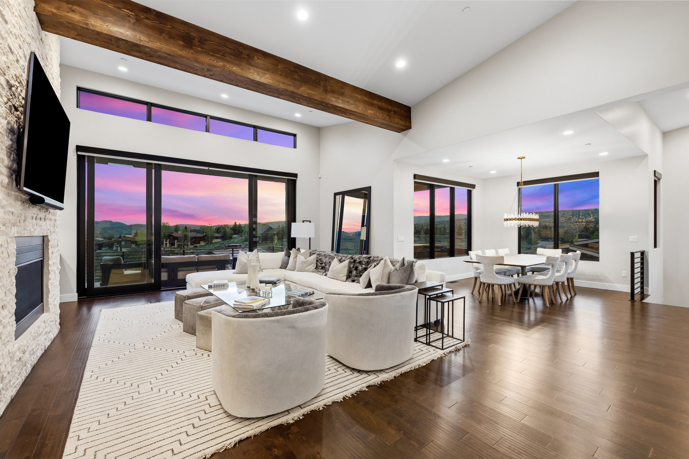 Expansive living area featuring soaring vaulted ceilings with exposed wood beams, wood-finish flooring, a stone-clad fireplace, and full-height glass doors