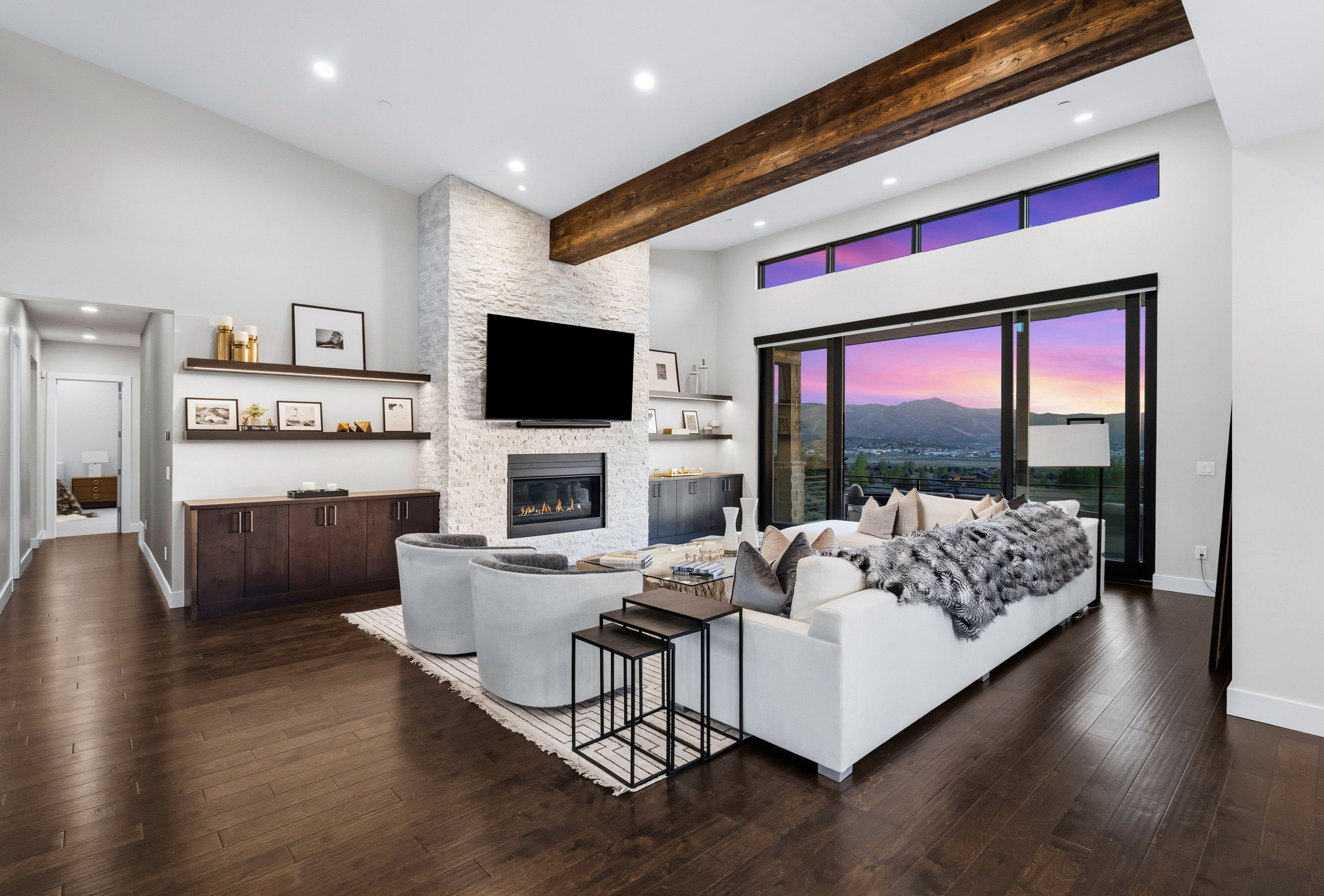 Spacious living area featuring high ceilings with exposed wood beams, a stone-clad fireplace, built-in shelving, dark wood-finish flooring, and large sliding glass doors overlooking distant mountains