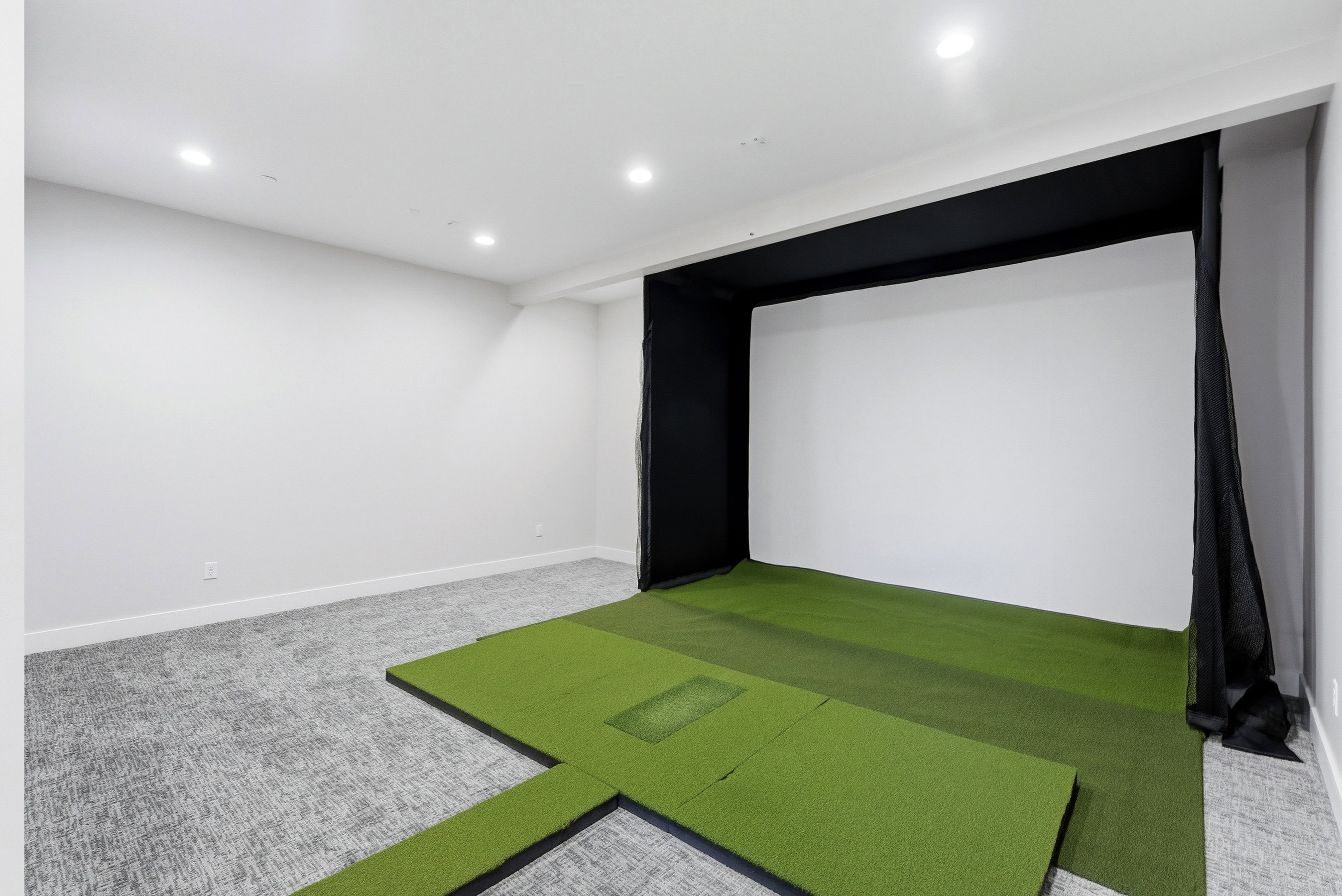 Dedicated indoor golf simulator space featuring a projection screen, artificial turf flooring, recessed lighting, white walls, and patterned carpet