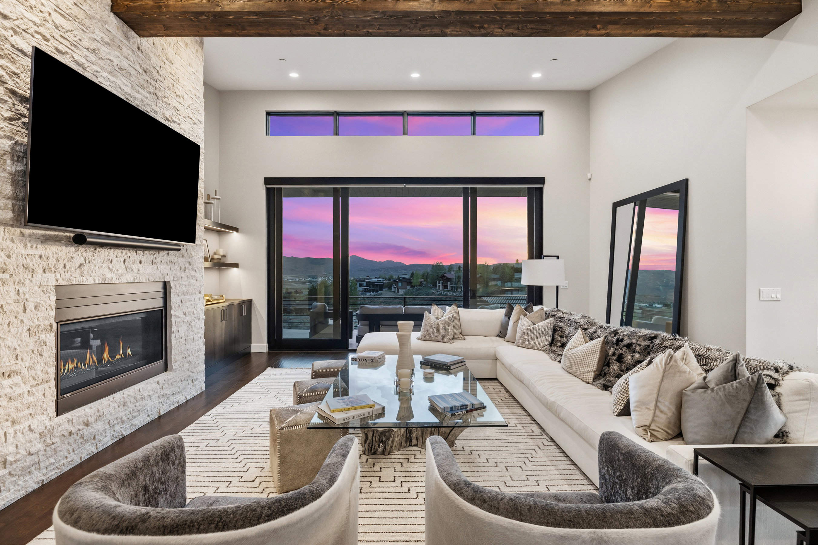 Spacious living area featuring high ceilings, exposed wood beams, a stone-clad fireplace, dark wood-finish flooring, and expansive glass doors opening to the outdoors