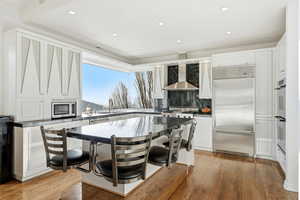Kitchen featuring white cabinets, built in appliances, wall chimney exhaust hood, a breakfast bar area, and light wood-style flooring