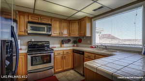 Kitchen with stainless steel appliances, tile counters, brown cabinets, and a drop ceiling
