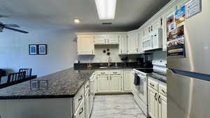 Kitchen featuring white appliances, a peninsula, dark stone countertops, white cabinets, and light wood-style flooring