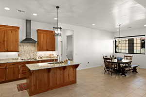 Kitchen with brown cabinets, a chandelier, wall chimney range hood, light stone countertops, and recessed lighting