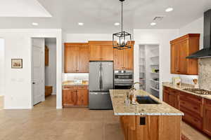 Kitchen featuring stainless steel appliances, brown cabinetry, wall chimney range hood, light stone countertops, and recessed lighting
