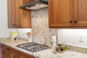 Kitchen with brown cabinets, under cabinet range hood, light stone countertops, and stainless steel gas stovetop