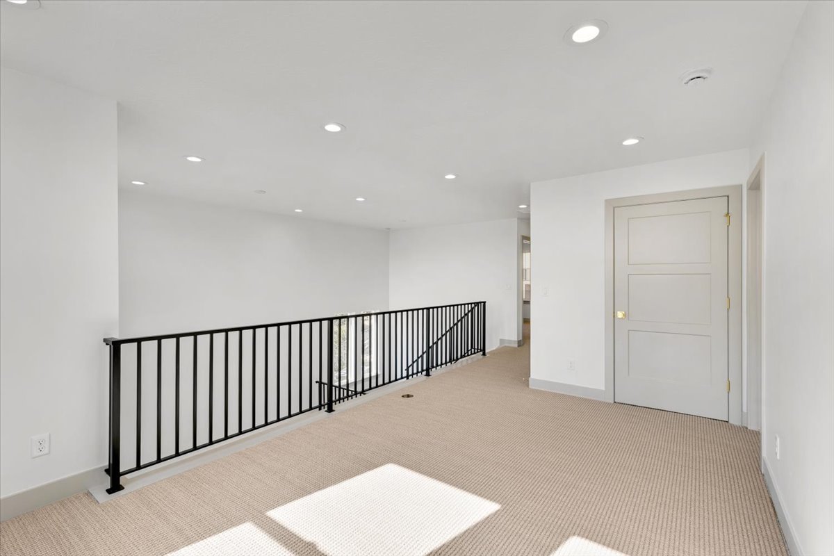 Spacious upper-level landing featuring neutral carpeting, recessed ceiling lighting, a modern black metal railing, and a contemporary three-panel interior door