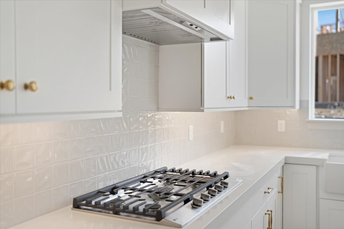 Gourmet kitchen featuring a built-in gas range, coordinating range hood, white shaker cabinetry with gold-tone hardware, textured tile backsplash, and light-toned countertops