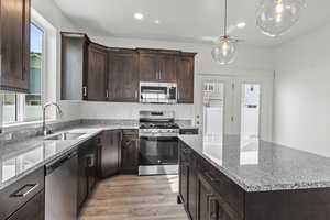 Kitchen featuring dark brown cabinetry, appliances with stainless steel finishes, light stone counters, hanging light fixtures, and light wood-style floors