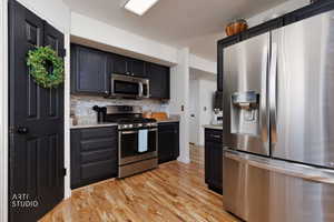 Kitchen with stainless steel appliances, light stone countertops, backsplash, dark cabinets, and light wood-type flooring