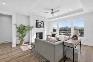 Living room featuring ceiling fan, a mountain view, a tray ceiling, light wood finished floors, and a glass covered fireplace