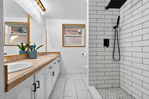 Full bath featuring double vanity, tiled shower, light marble finish flooring, and a textured ceiling