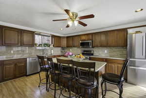 Kitchen with appliances with stainless steel finishes, brown cabinetry, light stone counters, light wood-style flooring, and recessed lighting
